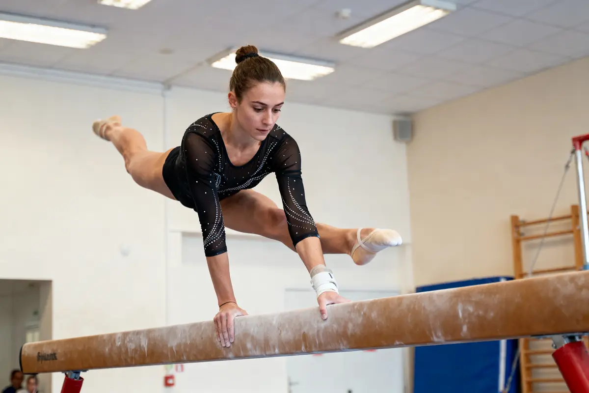 Gymnaste en justaucorps noir exécutant un équilibre sur poutre dans une salle de sport.