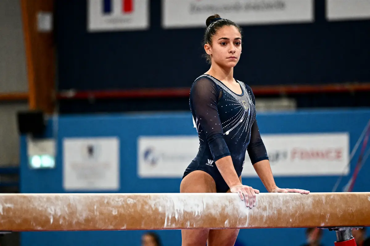 Jeune gymnaste en justaucorps noir concentrée sur une poutre, fond avec drapeau français flou.