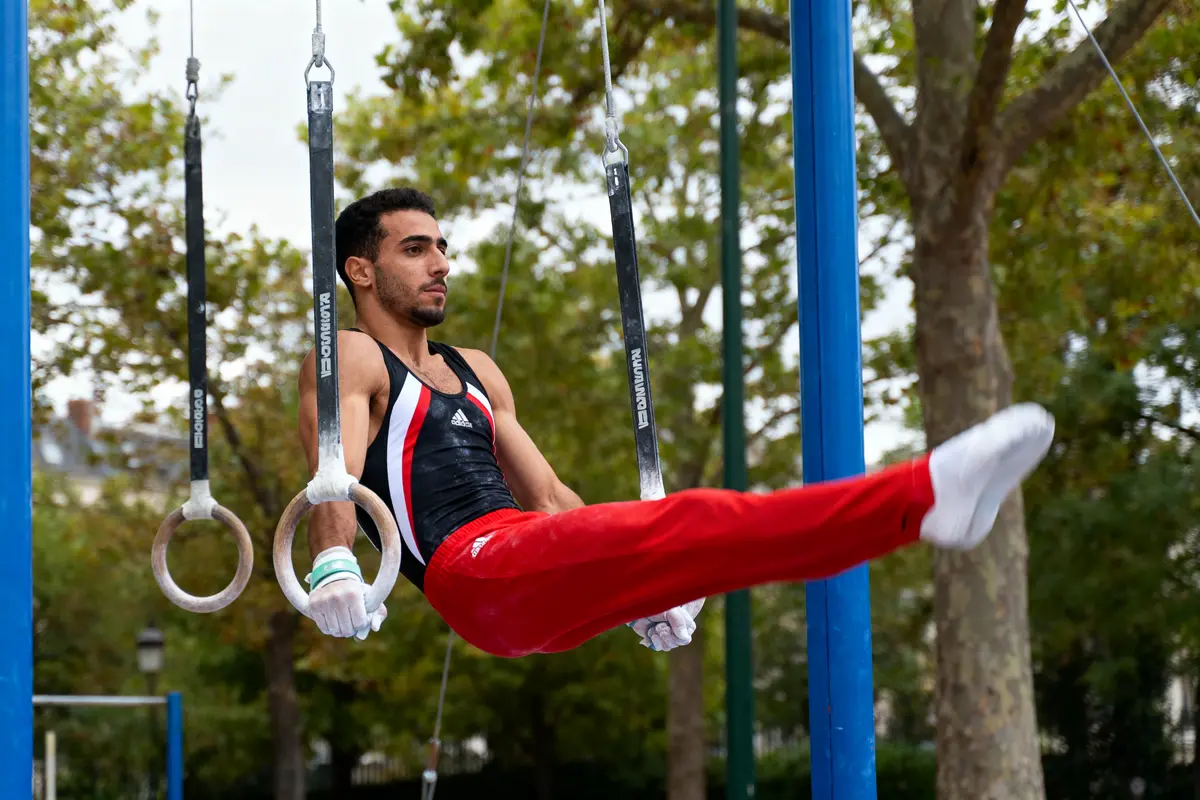 Gymnaste en tenue rouge effectuant une figure sur anneaux en extérieur, entouré d'arbres.