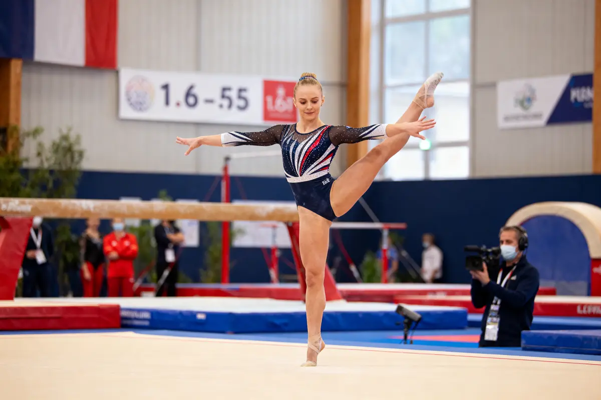 Gymnaste exécutant un mouvement de danse sur le sol, en compétition, portant un justaucorps bleu et rouge.