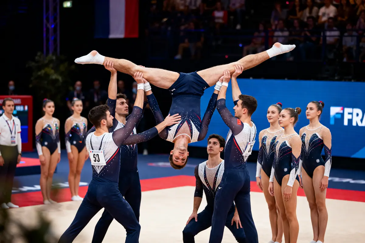 Groupe de gymnastes en uniforme effectuant une figure acrobatique synchronisée sur un tapis, drapeau français visible.