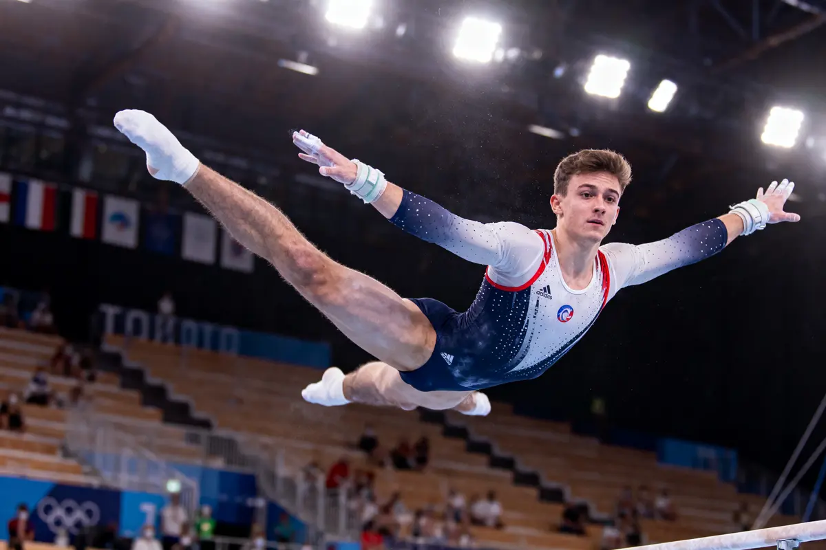 Gymnaste en plein saut, bras tendus, sur une poutre, lors d'une compétition olympique.