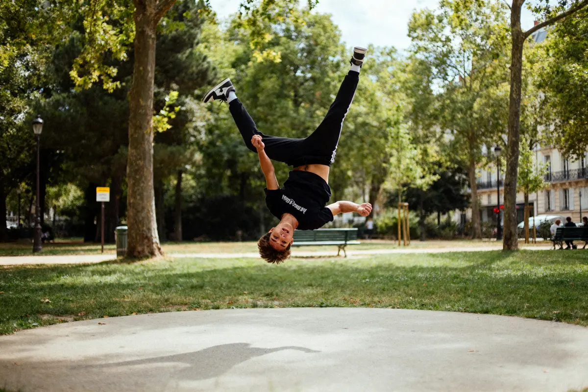Jeune homme exécutant un flip à l'envers dans un parc verdoyant, entouré d'arbres et de pelouses.